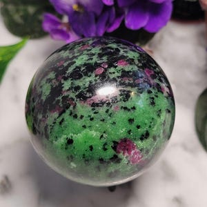 May include: A polished sphere of ruby in zoisite. The gemstone features a vibrant green base speckled with black and flecks of ruby red. The sphere is smooth and reflective, with a glossy finish. The background includes purple flowers.