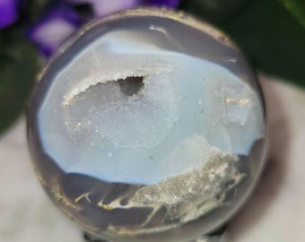 May include: A polished spherical stone featuring a translucent grey and white banded agate exterior with a central geode cavity lined with sparkling white druzy crystals. The smooth surface displays natural mineral inclusions and light reflections.