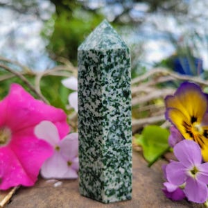 May include: A green and white speckled stone obelisk stands upright, surrounded by pink, purple, and yellow flowers. The obelisk has a pointed top and a rectangular base. The background is blurred, with green foliage and a blue sky.