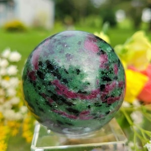 May include: A polished spherical stone featuring a mottled pattern of deep green, vibrant magenta, and black mineral inclusions. The surface is smooth and reflective, showcasing a natural blend of colors throughout the rounded gemstone form.