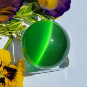 May include: A glossy green sphere with a cat's eye effect, displayed on a clear glass stand. The sphere is set against a backdrop of yellow and purple pansies and green leaves. The sphere is a decorative item.