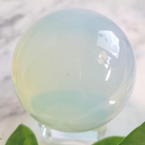 May include: A translucent, opalescent sphere, possibly a crystal ball, with a soft gradient of colors from pale blue to white. The sphere is resting on a clear stand, with green leaves in the foreground.