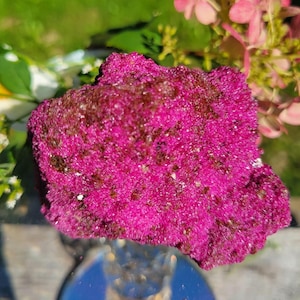 May include: A vibrant magenta mineral specimen with a crystalline, textured surface featuring dark brown inclusions. The rock displays a dense, needle-like structure across its irregular, jagged shape, reflecting light with a subtle, sparkling sheen.
