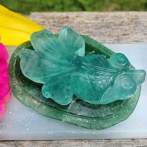 May include: A translucent, light green carved gemstone figurine shaped like a goldfish with detailed fins and scales, resting on a dark green, textured, oval-shaped stone base. The carving features prominent, rounded eyes and a smooth, polished surface.