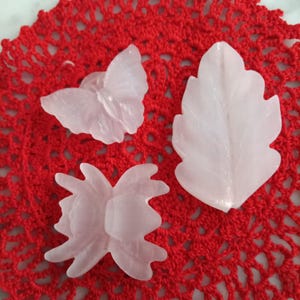 May include: Three white wax candles shaped like a butterfly, a leaf, and a beetle. The candles are on a red crocheted doily.