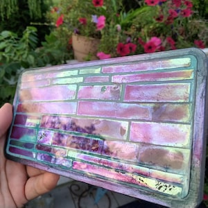 May include: A rectangular iridescent tile with a brick pattern. The tile is made of glass and has a pink, purple, and green color scheme.