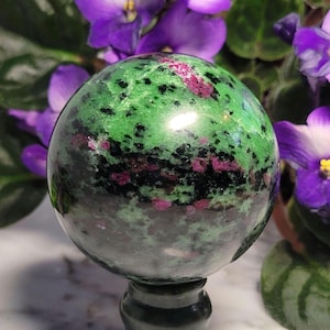 May include: A polished sphere of green zoisite with ruby inclusions, resting on a dark green stand. The gemstone features a mottled pattern of green, black, and pink, with a glossy finish. Purple flowers are visible in the background.