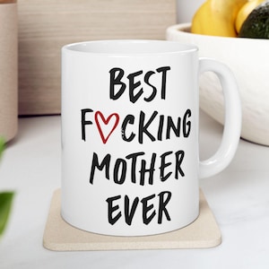 May include: White ceramic mug with the text "BEST F*CKING MOTHER EVER" in black lettering. The heart in "F*CKING" is red. The mug sits on a beige coaster.