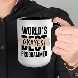 Puede incluir: Taza de café blanca con borde y asa negros. La taza tiene texto negro que dice "WORLD'S OKAYEST BEST PROGRAMMER" con la palabra "BEST" parcialmente cubierta por una etiqueta de cinta marrón que dice "OKAYEST".
