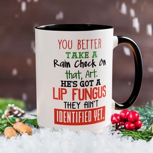May include: White coffee mug with a black rim and handle. The mug has a humorous saying printed on it in red, green, and black text: "You better take a rain check on that, Art. He's got a lip fungus they ain't identified yet."