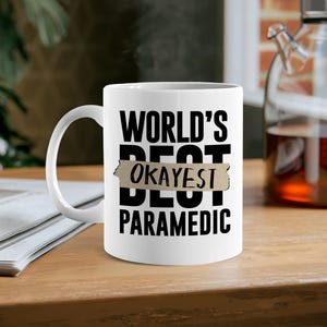 May include: White ceramic mug with the text "WORLD'S BEST OKAYEST PARAMEDIC" in black lettering. The word "BEST" is partially covered with a tan-colored tape. The mug is on a wooden surface.