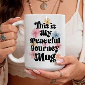 May include: A white ceramic mug featuring the black text This is My Peaceful Journey MUG in a bold, retro-style font. The text is surrounded by small, colourful illustrations of pink, yellow, and light blue flowers scattered around the lettering.