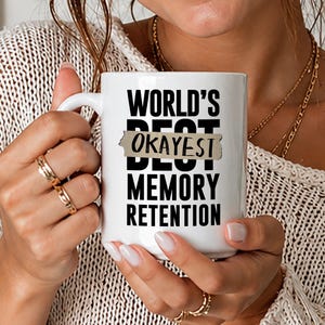 May include: White ceramic mug with the text "WORLD'S BEST OKAYEST MEMORY RETENTION" in black. The word "OKAYEST" is in a tan rectangle. The mug is held by a person wearing gold rings.