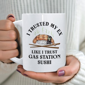 Puede incluir: Una taza de cerámica blanca con un gráfico de una pieza de sushi de camarón con líneas de vapor verdes encima, palillos cruzados debajo y el texto negro I TRUSTED MY EX LIKE I TRUST GAS STATION SUSHI en una fuente negrita sin remates.