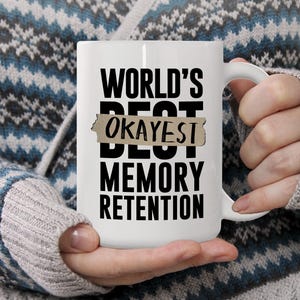 May include: White ceramic mug with the text "WORLD'S BEST OKAYEST MEMORY RETENTION" in black. The word "OKAYEST" is on a tan, rectangular background. The mug is held by a person wearing a blue and white patterned sweater.