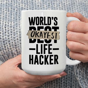 May include: White ceramic mug with the text "WORLD'S BEST OKAYEST LIFE HACKER" in black. The word "BEST" is partially covered with a tan-colored rectangle. The mug has a large handle.
