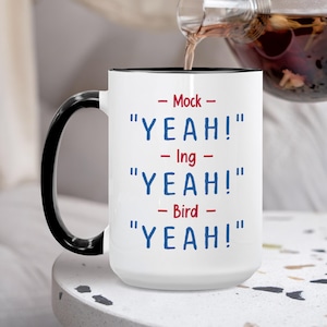 May include: White ceramic mug with a black handle and rim. The mug features the text "- Mock - YEAH! - Ing - YEAH! - Bird - YEAH!" in blue and red font. Tea is being poured into the mug.