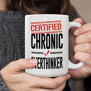 May include: A white ceramic mug featuring the red and black text CERTIFIED CHRONIC OVERTHINKER with a red checkmark symbol between the words CHRONIC and OVERTHINKER. The design includes horizontal red and black lines above and below the text.
