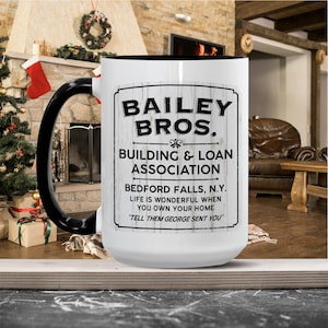 Könnte beinhalten: Eine weiße Keramiktasse mit schwarzem Henkel und Rand. Die Tasse zeigt den Text "BAILEY BROS. BUILDING & LOAN ASSOCIATION BEDFORD FALLS, N.Y." und das Zitat "TELL THEM GEORGE SENT YOU."
