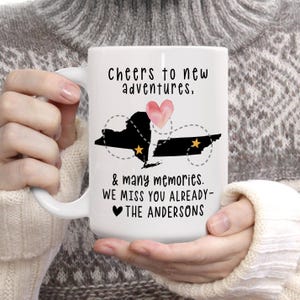 May include: White ceramic mug with the text "Cheers to new adventures, & many memories. We miss you already - The Andersons." The mug features black silhouettes of two states, a pink heart, and gold stars.