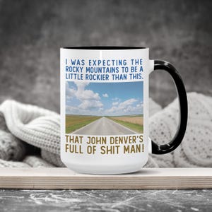 May include: White ceramic mug with a black handle and rim. The mug features a landscape image of a road under a blue sky and text that reads, "I WAS EXPECTING THE ROCKY MOUNTAINS TO BE A LITTLE ROCKIER THAN THIS. THAT JOHN DENVER'S FULL OF SHIT MAN!"