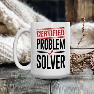 May include: White ceramic mug with the text "CERTIFIED PROBLEM SOLVER" in red and black. The mug has a large handle and is set against a blurred background of wood and a cream-colored knitted item.