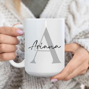 Op de afbeelding: Witte keramische mok met een grote grijze letter "A" en de naam "Ariana" in een sierlijk lettertype. De mok heeft een gebogen handvat. De mok wordt vastgehouden door een persoon die een witte trui draagt.