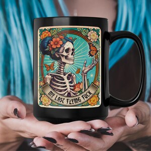 La mia ultima tazza volante da scopare, una carta in stile tarocchi con una donna scheletro che guarda una farfalla, un design gotico colorato su una tazza di ceramica nera