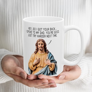 Peut inclure: Un mug en céramique blanche orné d'une illustration représentant une figure aux longs cheveux bruns et à la barbe, vêtue d'une tunique jaune et d'une robe bleue. Au-dessus, le texte noir indique : HEY...SO I GOT YOUR BACK... I SPOKE TO MY DAD. YOU'RE GOOD. JUST TRY HARDER NEXT TIME.