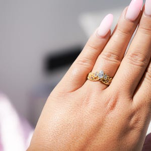 May include: A gold-colored ring with a crown design, featuring clear and red gemstones. The ring is worn on a finger, showcasing its intricate details and elegant style. The ring's design is reminiscent of a tiara.