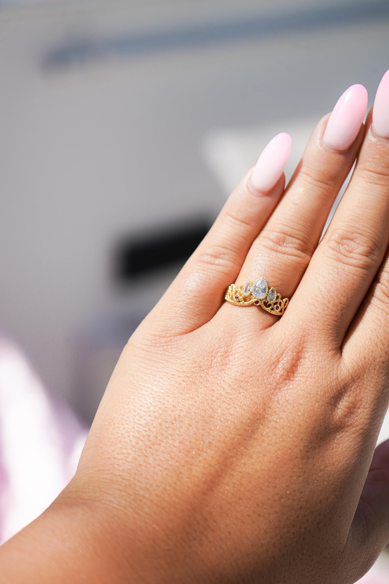 May include: A gold-colored ring with a crown design, featuring clear and red gemstones. The ring is worn on a finger, showcasing its intricate details and elegant style. The ring's design is reminiscent of a tiara.