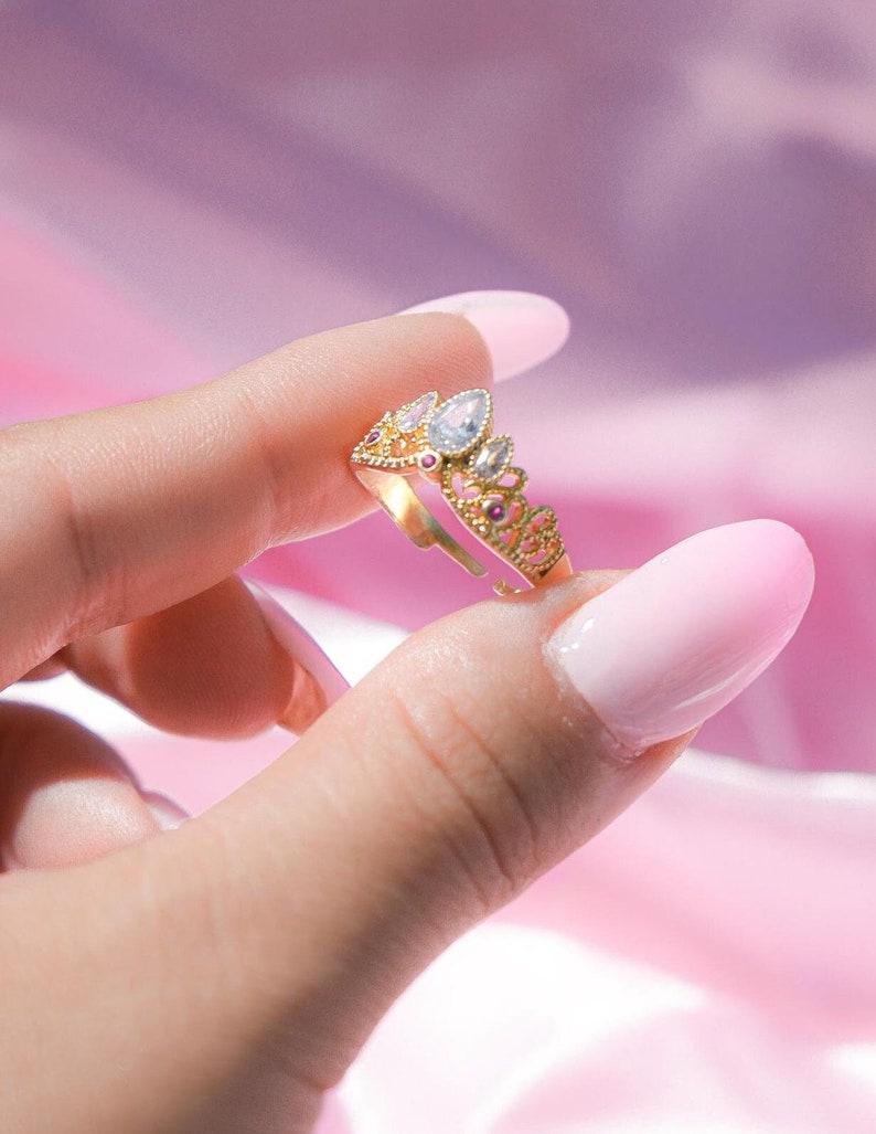 May include: A gold-colored crown ring with clear and pink gemstones. The ring has a delicate design with a teardrop-shaped center stone and smaller gems. The ring is held against a pink background.