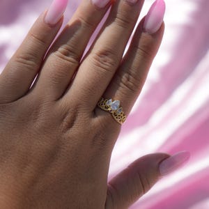 May include: A gold-colored ring with a crown design, featuring a pear-shaped clear gemstone. The ring is on a hand with long, oval-shaped nails painted in a pink ombre style. The background is a pink, silky fabric.