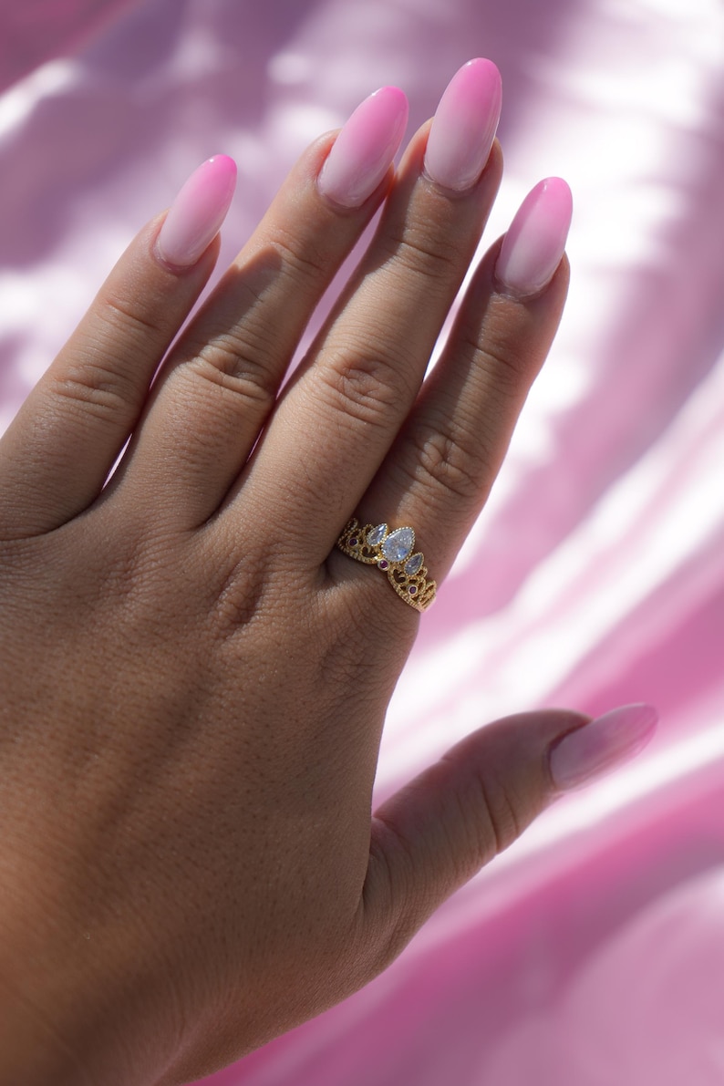 May include: A gold-colored ring with a crown design, featuring a pear-shaped clear gemstone. The ring is on a hand with long, oval-shaped nails painted in a pink ombre style. The background is a pink, silky fabric.