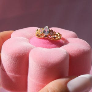 May include: A gold-colored ring with a crown design, featuring a pear-shaped, light blue gemstone at the center, flanked by smaller gems. The ring is displayed on a pink, flower-shaped jewelry box.