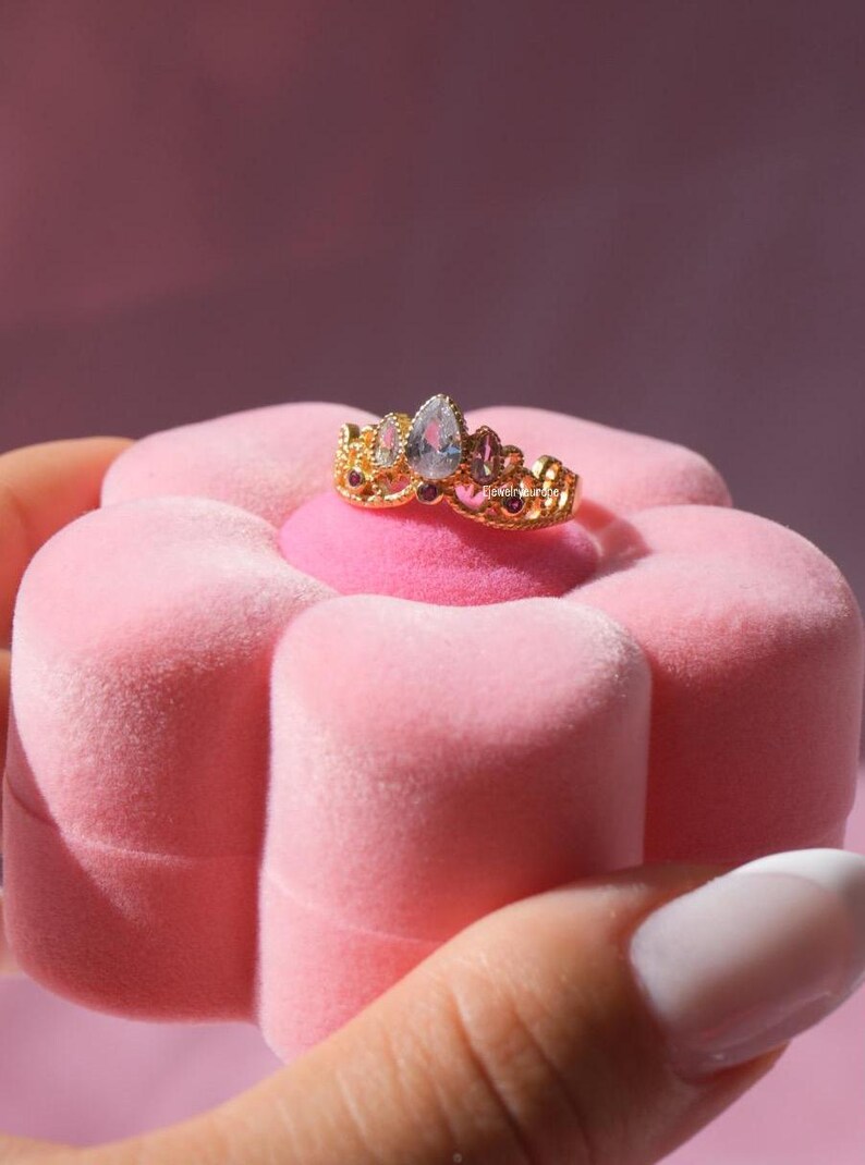 May include: A gold-colored ring with a crown design, featuring a pear-shaped, light blue gemstone at the center, flanked by smaller gems. The ring is displayed on a pink, flower-shaped jewelry box.