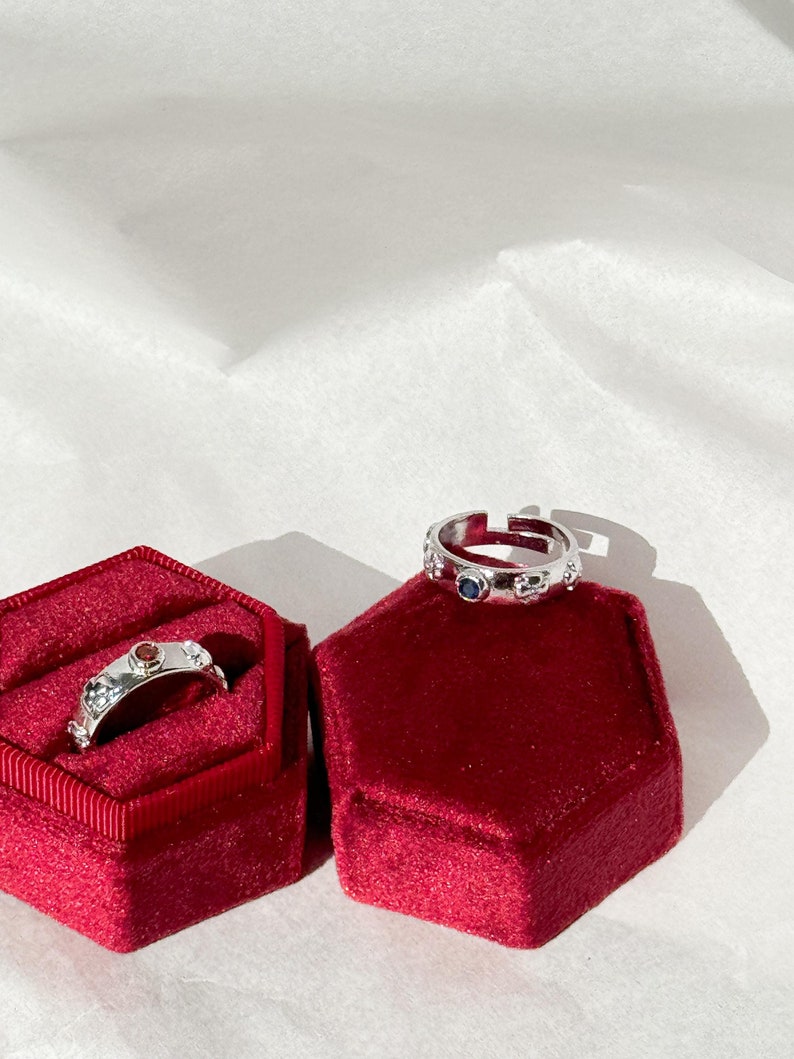 May include: Two silver rings with colorful gemstones set in red velvet ring boxes. The ring on the left has a red gemstone and the ring on the right has a blue gemstone.
