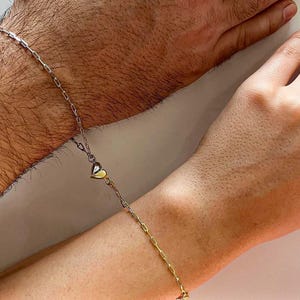 May include: Two silver chain bracelets connected by a gold heart charm. The bracelets are worn on the wrists of two people.