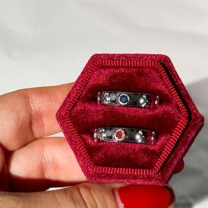 May include: Three silver rings with colorful gemstones in a red velvet ring box. The rings are stacked on top of each other. The top ring has a blue gemstone, the middle ring has a red gemstone, and the bottom ring has a clear gemstone.