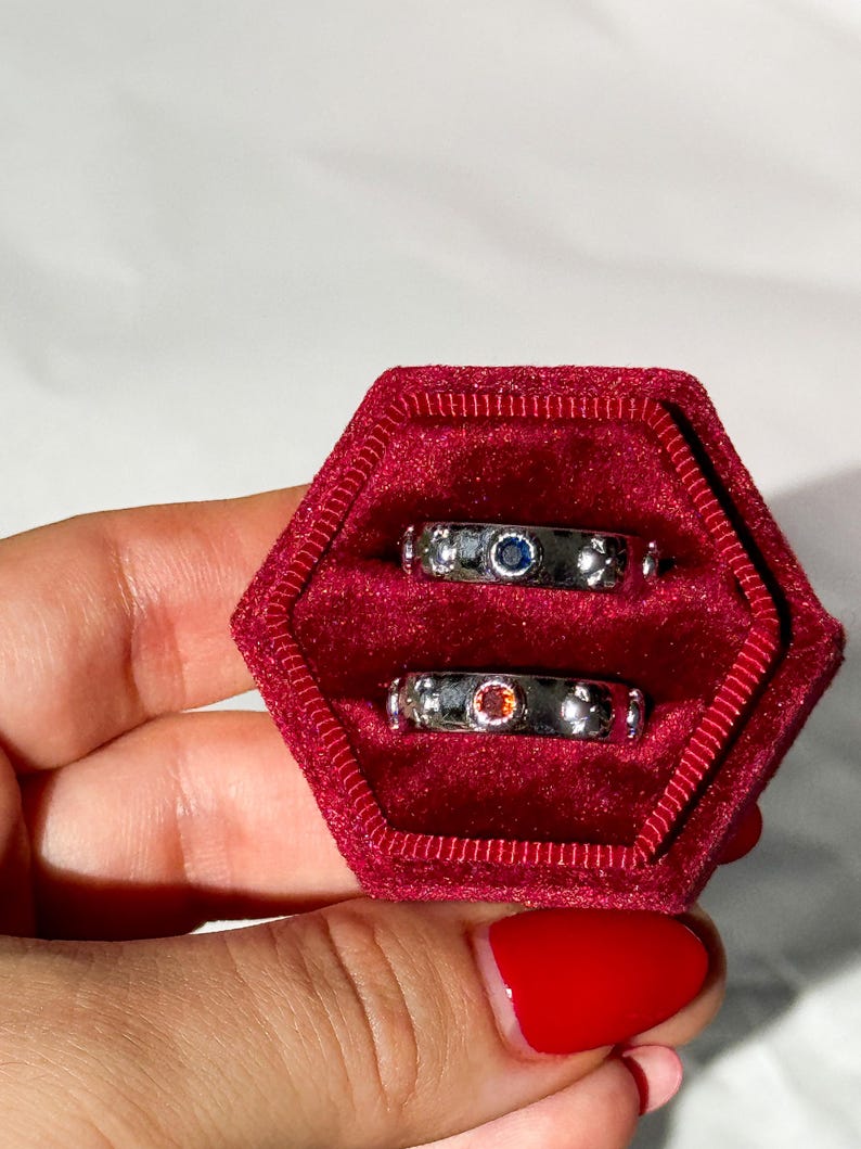 May include: Three silver rings with colorful gemstones in a red velvet ring box. The rings are stacked on top of each other. The top ring has a blue gemstone, the middle ring has a red gemstone, and the bottom ring has a clear gemstone.