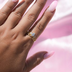 May include: A gold-colored crown ring with clear and red gemstones. The ring is on a hand with long, pink, oval-shaped nails. The background is a soft pink fabric.