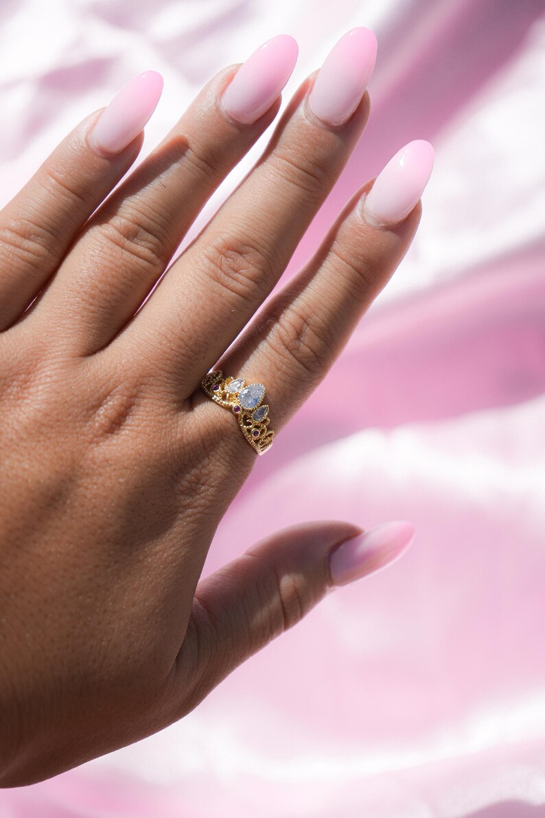 May include: A gold-colored crown ring with clear and red gemstones. The ring is on a hand with long, pink, oval-shaped nails. The background is a soft pink fabric.