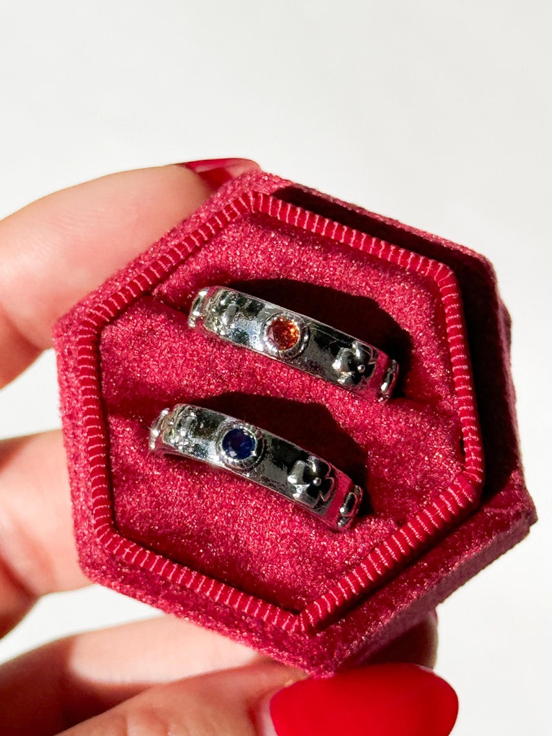 May include: Two silver rings with floral designs and small gemstones. The ring on top has a red gemstone, and the ring on the bottom has a blue gemstone. The rings are in a red velvet ring box.