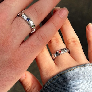 Peut inclure: Deux bagues en argent avec de petites pierres précieuses colorées. La bague sur la main gauche a une pierre bleue, et la bague sur la main droite a une pierre rouge.