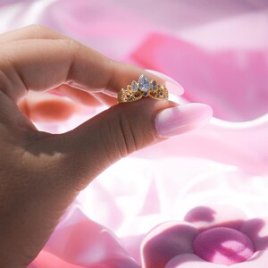 May include: A gold-colored ring in the shape of a crown, held between fingers. The ring features a central, pear-shaped, clear gemstone, flanked by smaller, teardrop-shaped gems and red accents. The background is a soft pink.