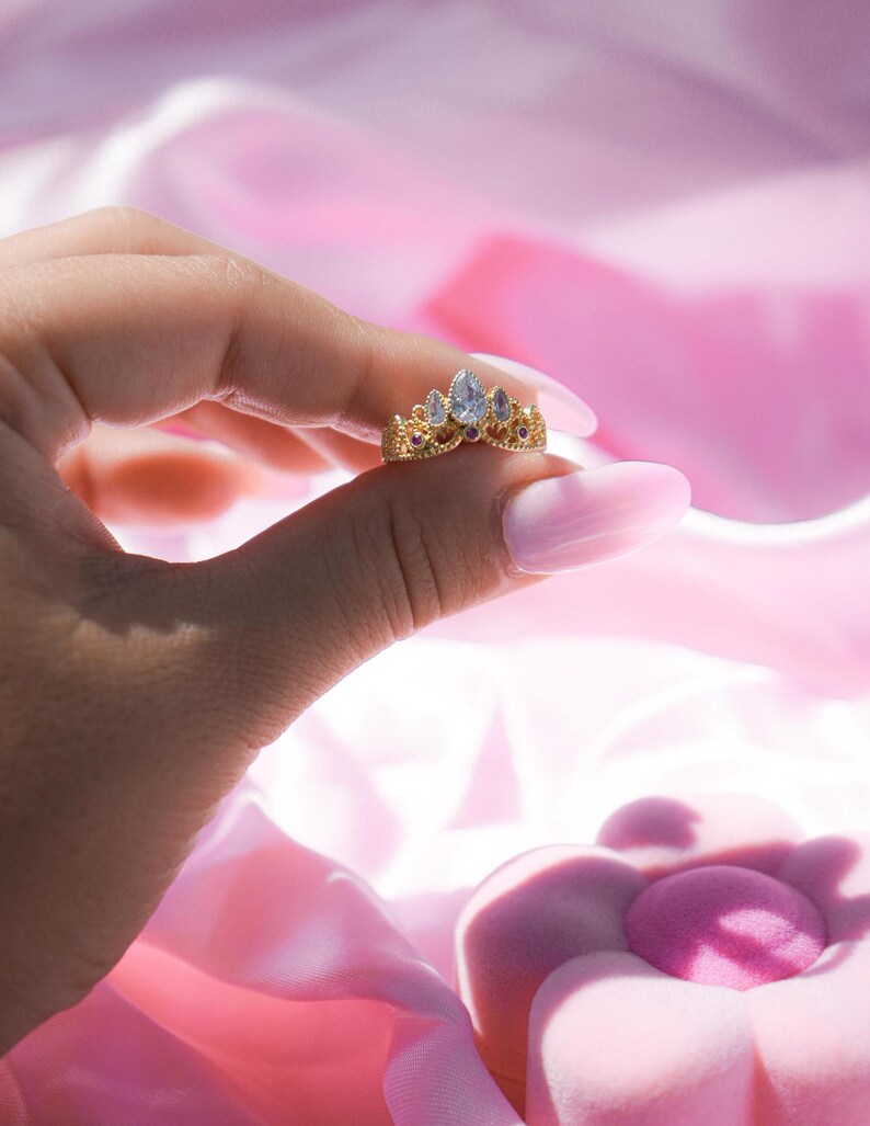May include: A gold-colored ring in the shape of a crown, held between fingers. The ring features a central, pear-shaped, clear gemstone, flanked by smaller, teardrop-shaped gems and red accents. The background is a soft pink.