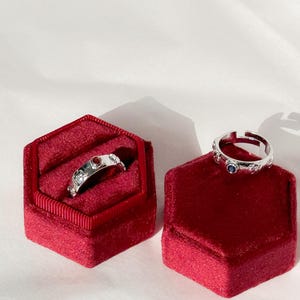 May include: Two silver rings with red and blue gemstones are displayed in red velvet ring boxes.
