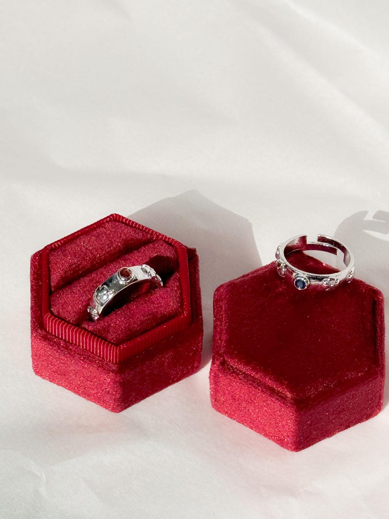 May include: Two silver rings with red and blue gemstones are displayed in red velvet ring boxes.