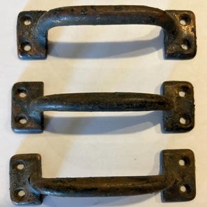 May include: Three antique bronze drawer pulls. Each pull has a curved handle and two rectangular mounting plates with two holes each. The bronze has a weathered, aged appearance with areas of discoloration and patina.