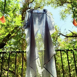 May include: A light blue sheer garment with a long, flowing design hangs on a hanger against a backdrop of green foliage. The garment has a subtle, faded look, suggesting it is vintage or handmade.